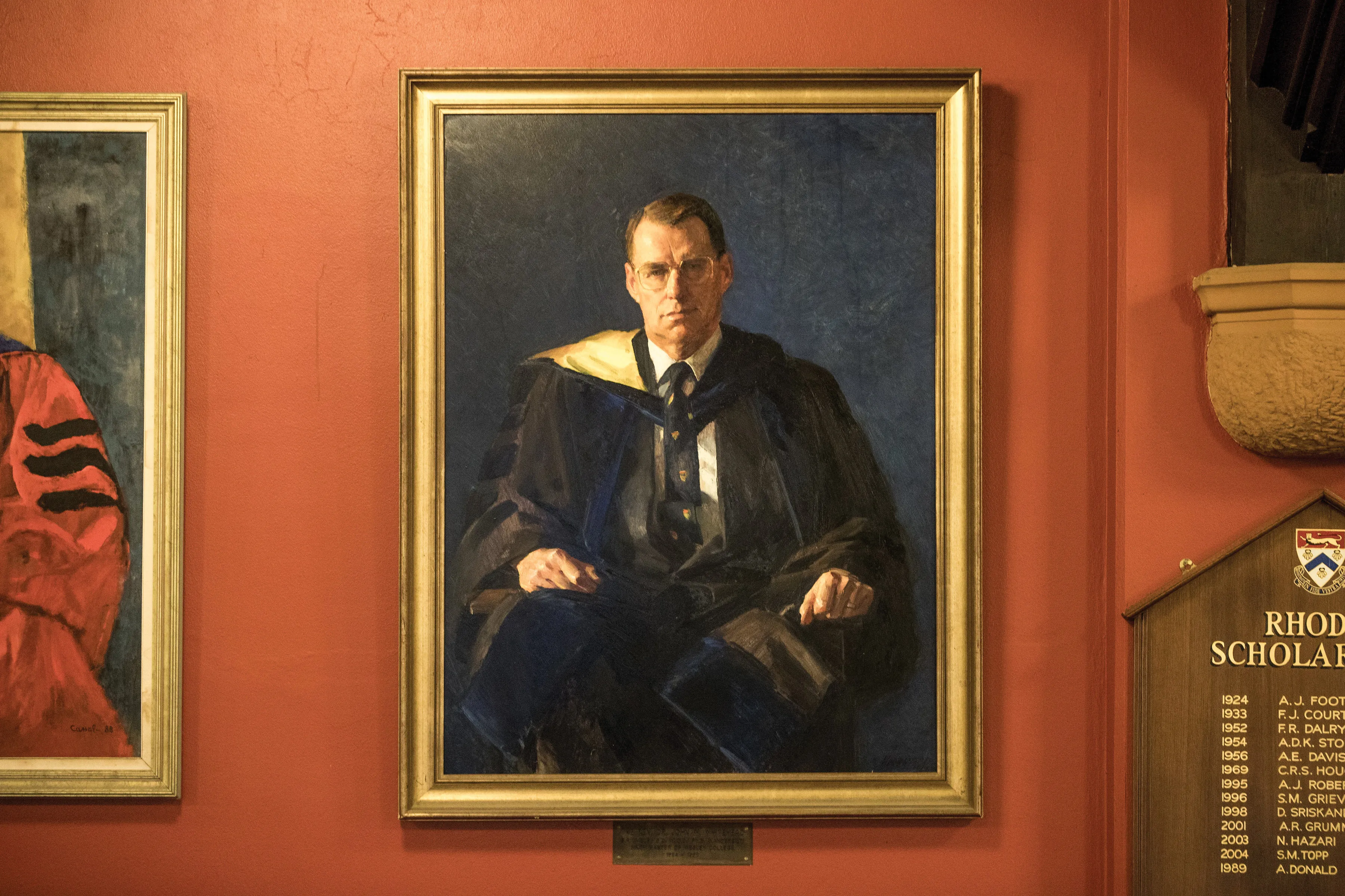 Framed portrait painting of a man wearing academic robes and glasses seated against a dark background on a red wall.