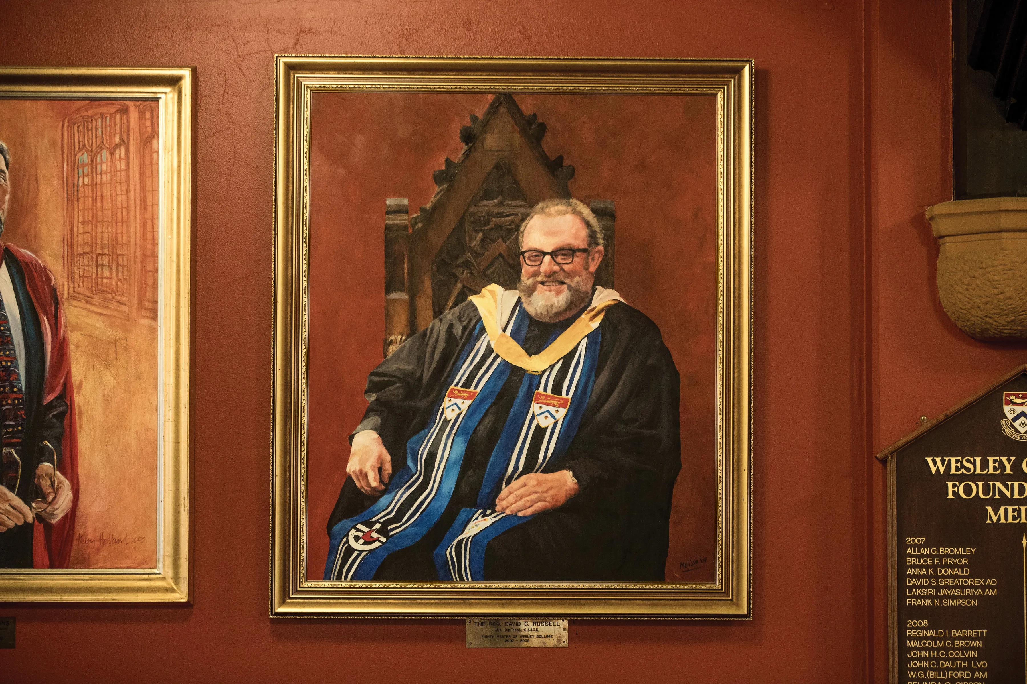 Portrait painting of a smiling man with glasses and a beard wearing academic regalia with a yellow-striped blue stole, seated on an ornate wooden chair with a red background.