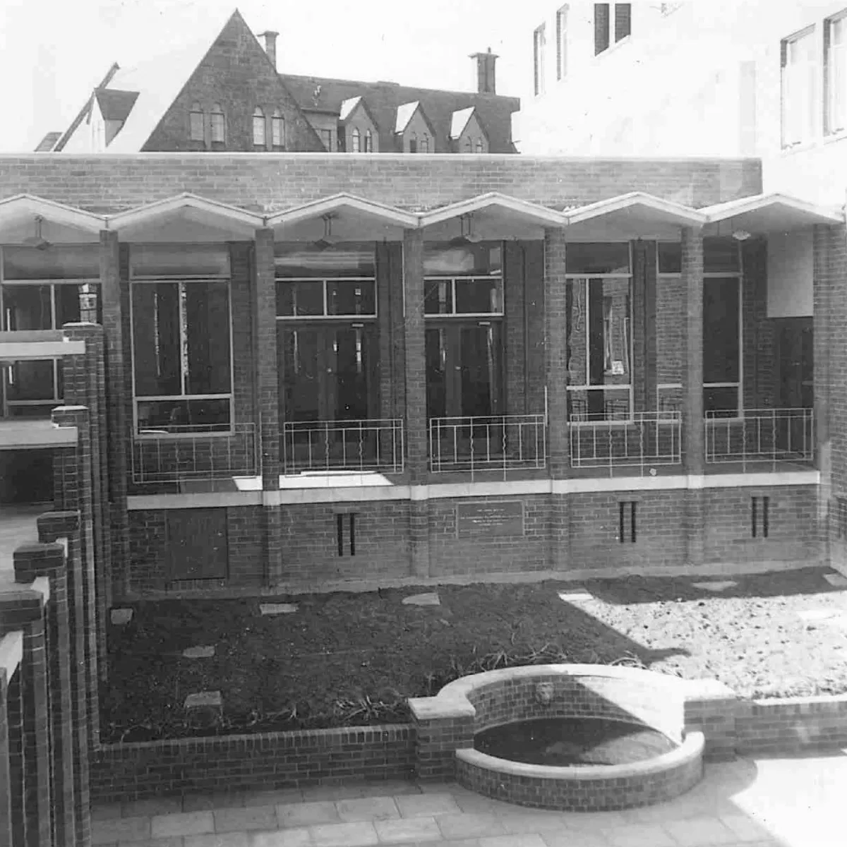 Black and white photo of a brick courtyard with raised garden beds and a rounded brick water feature, flanked by a building with large windows and a decorative roofline.