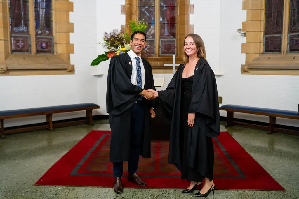 Two university graduates in academic gowns shaking hands inside a stained glass chapel.
