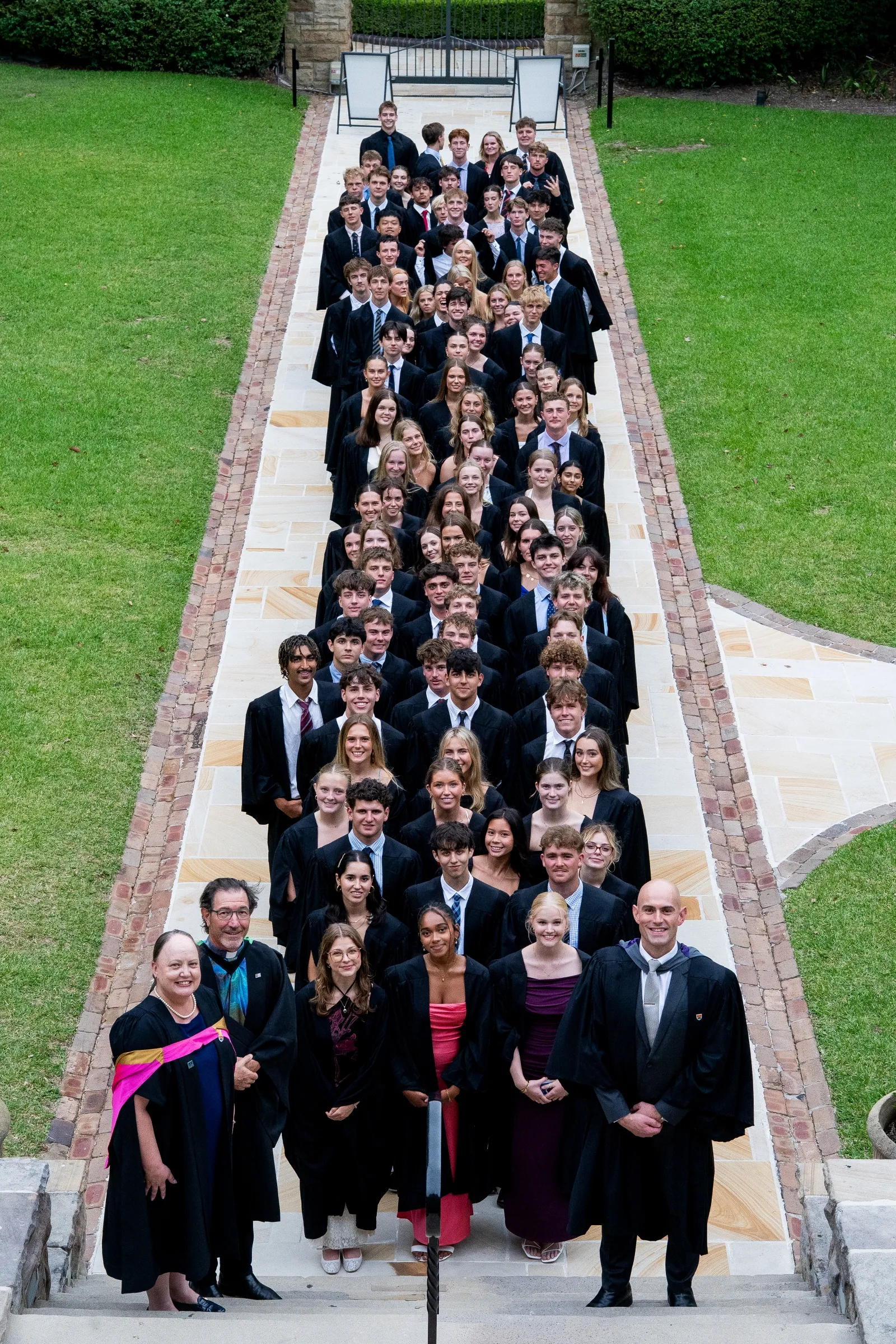 Group of graduating students and faculty in academic gowns posing on an outdoor walkway with grass on both sides.