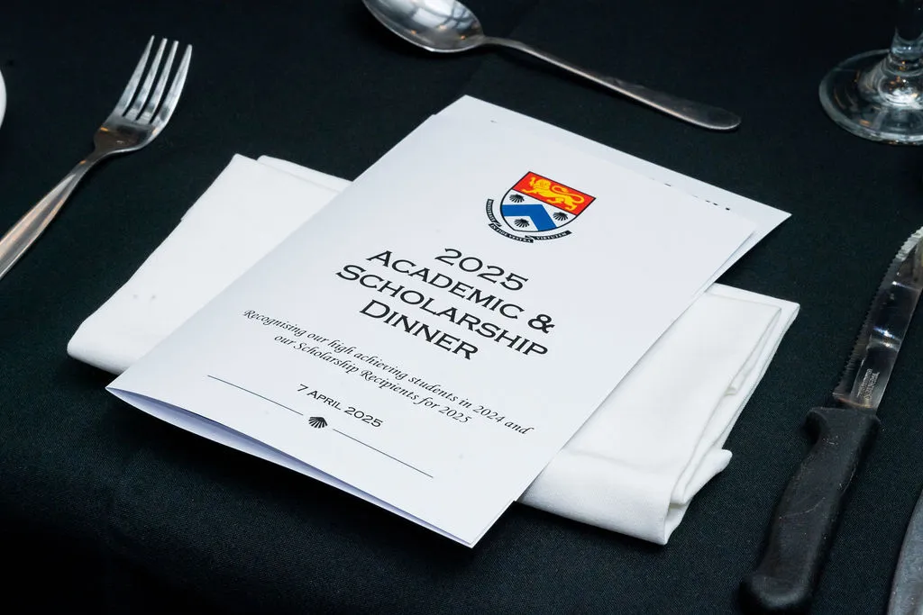 Program booklet for the 2025 Academic & Scholarship Dinner placed on a folded white napkin on a black tablecloth, with silverware around.