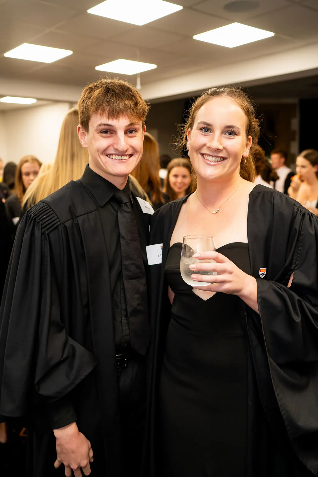 Two smiling young adults wearing black academic gowns at an indoor graduation event, one holding a glass of water.