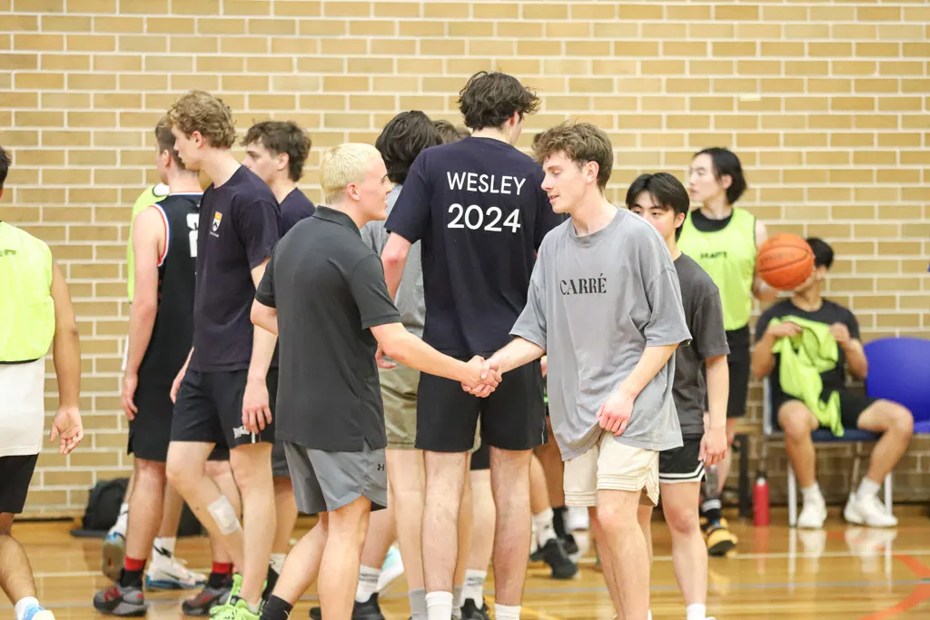 Young men in sportswear shaking hands on an indoor basketball court with a brick wall background.