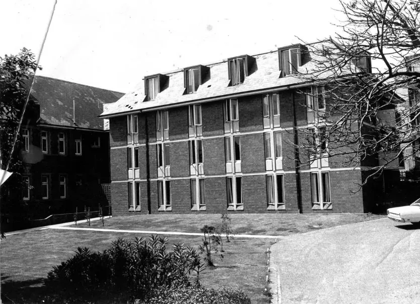 Black and white photo of a three-story brick residential building with dormer windows on the roof and a lawn in front.