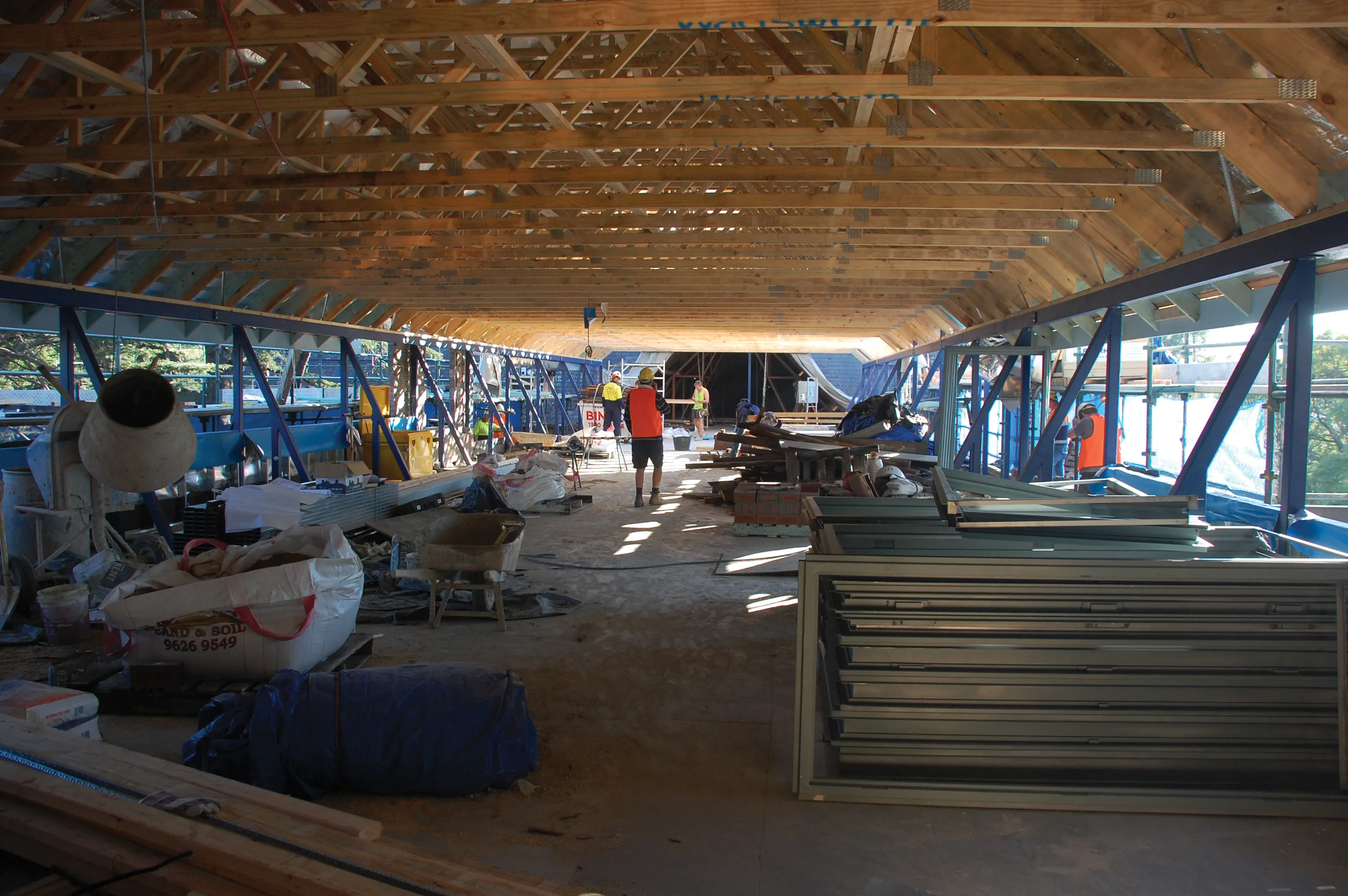 Construction site inside a building with wooden roof trusses, workers, metal materials, and construction equipment.