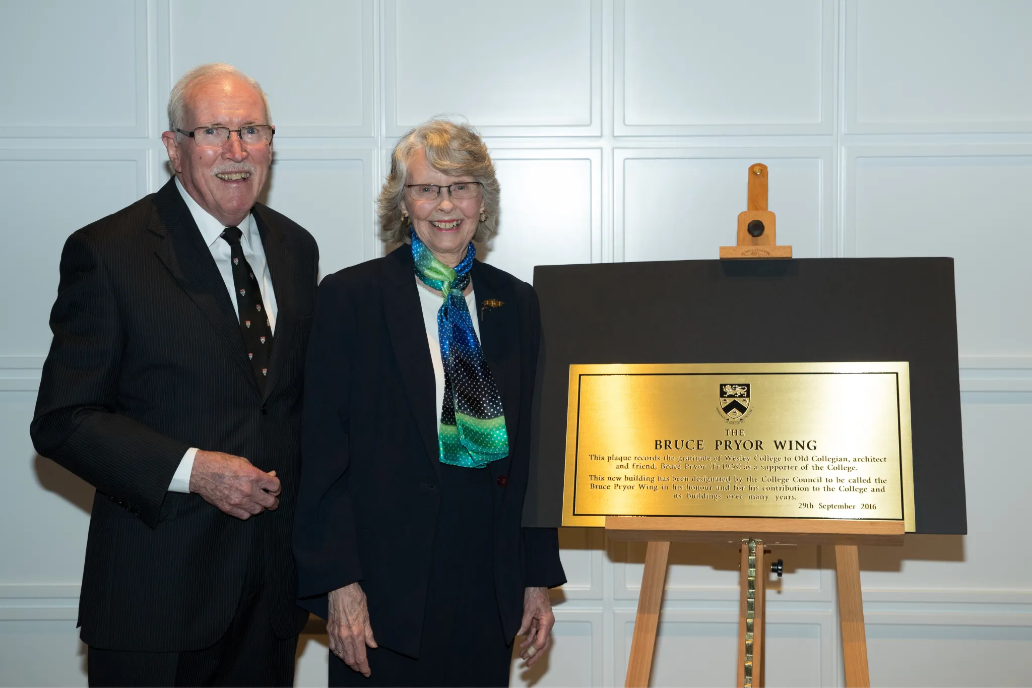 An elderly man and woman in formal attire standing next to a plaque on an easel reading 'The Bruce Pryor Wing' at a commemorative event.