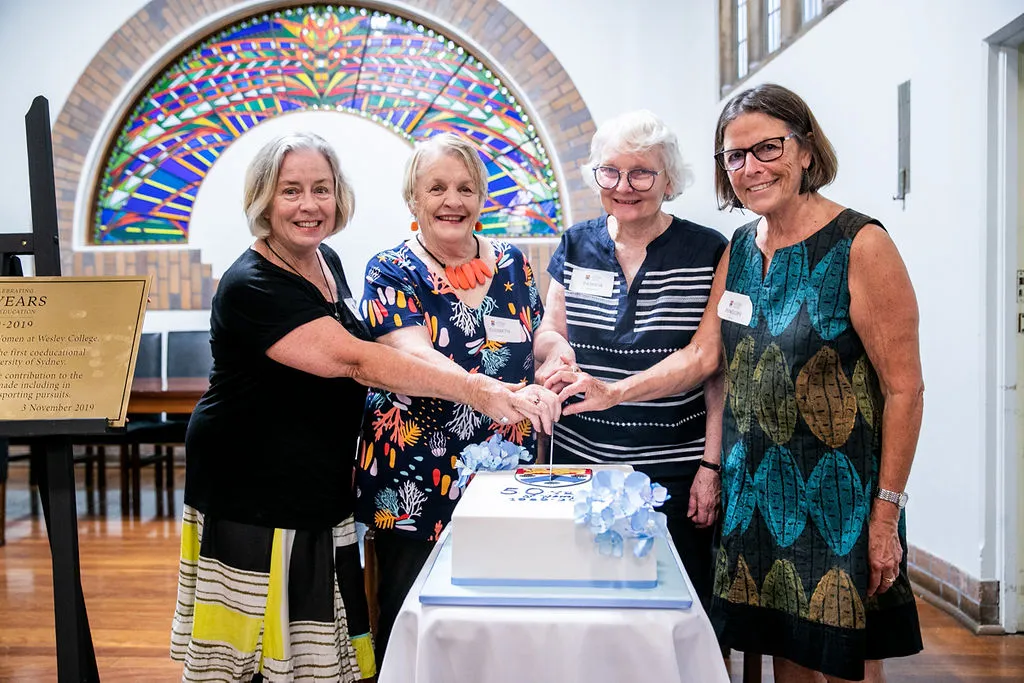 Four women standing together cutting a decorated cake on a white table in a room with a colorful stained glass arch in the background.
