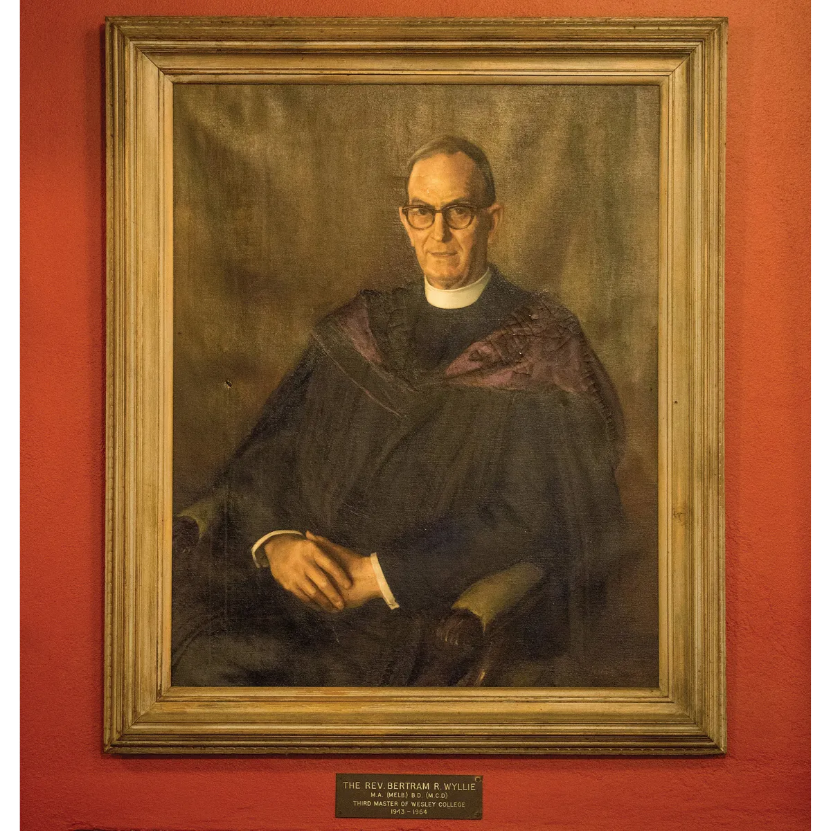 Framed oil painting of a man wearing glasses and clerical collar seated in a chair against a brown background.