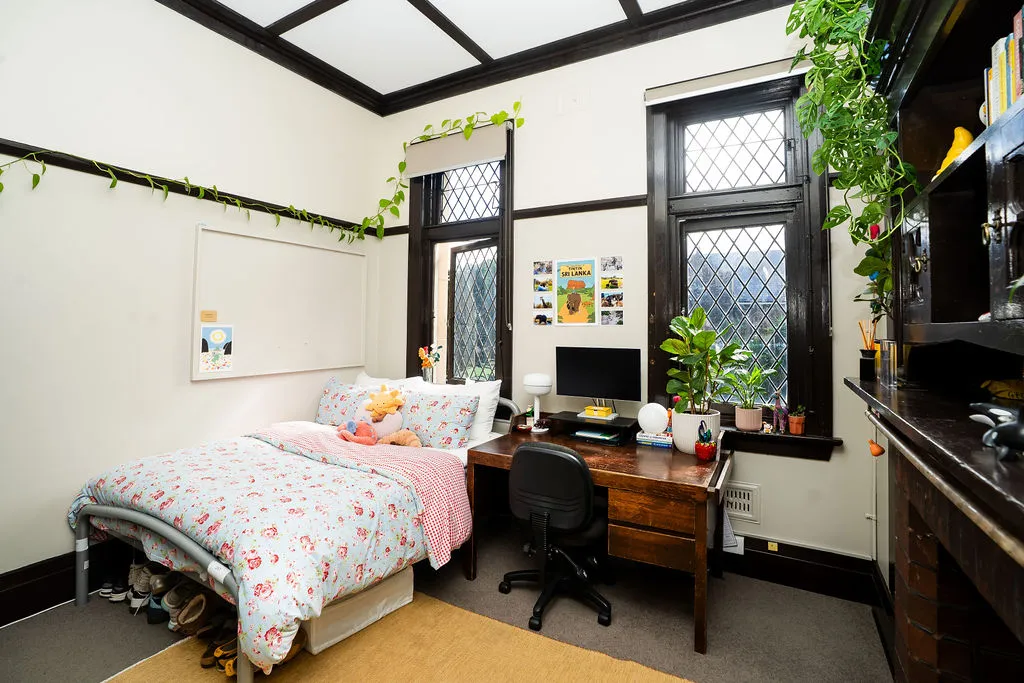 Cozy bedroom with a floral bedspread, stuffed toys on the bed, a wooden desk with a computer, potted plants, and lattice windows with dark wood trim.