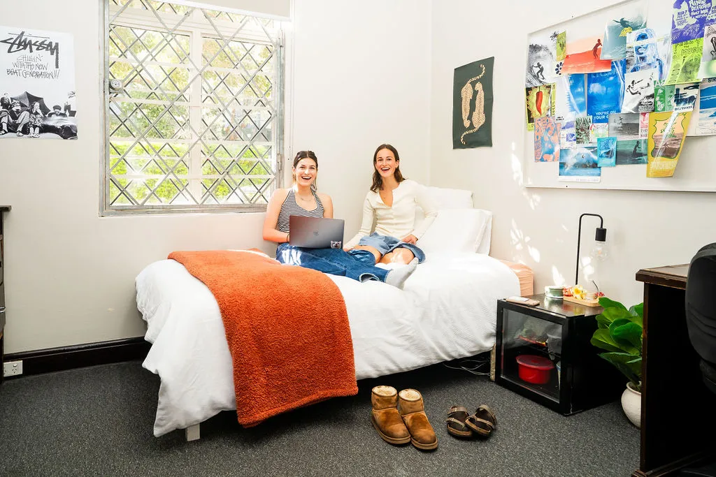Two women sitting on a bed with a laptop in a bright, cozy bedroom featuring a white bedspread, orange throw, and a bulletin board with colorful pictures on the wall.