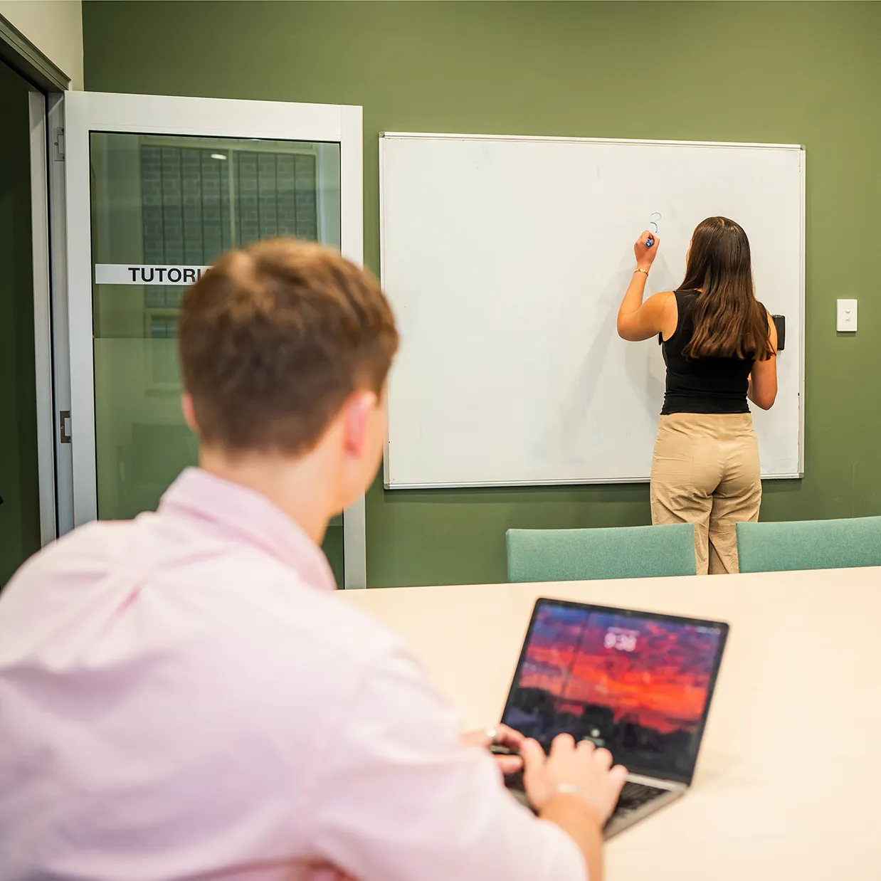 A woman writes a question mark on a whiteboard while a man in a pink shirt uses a laptop in a tutoring room.