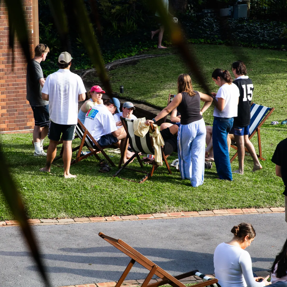 Group of people socializing outdoors on grass with striped deck chairs, some standing and some sitting.