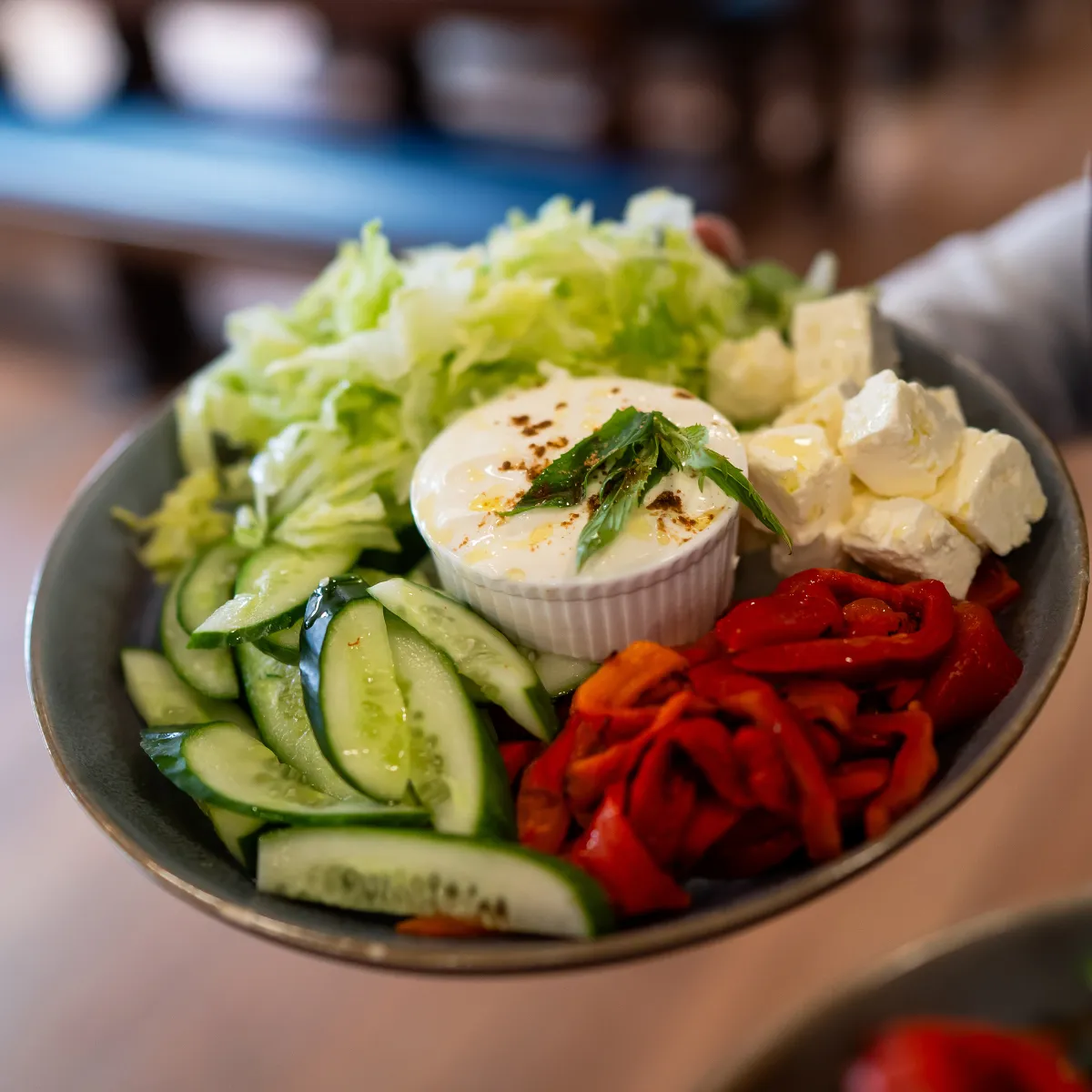 Plate with fresh cucumber slices, lettuce, roasted red peppers, cubes of feta cheese, and a small bowl of creamy dip garnished with herbs.