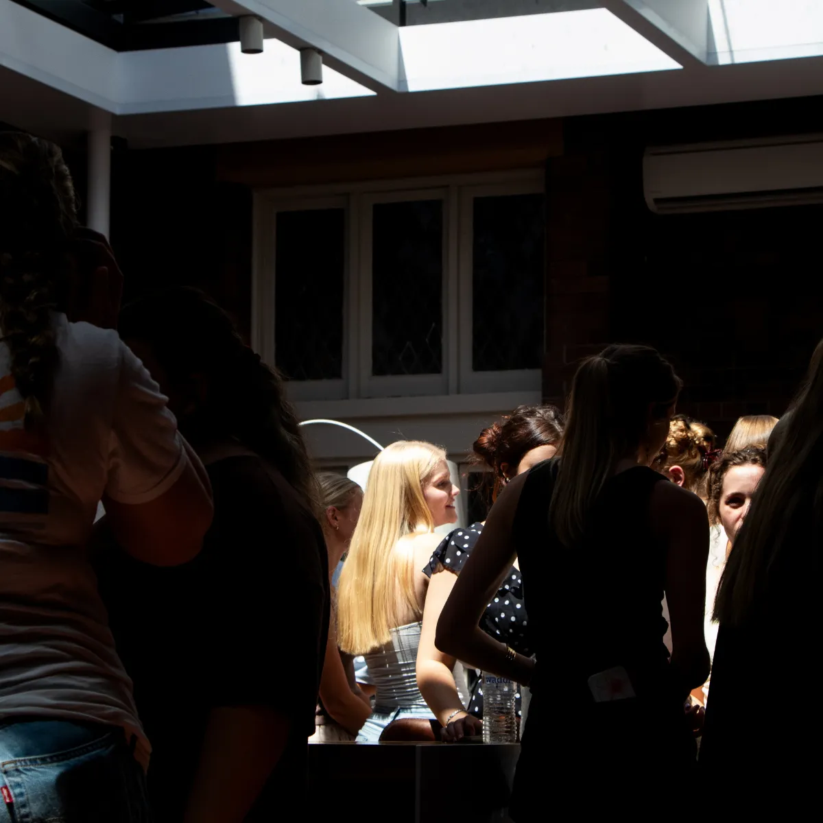 Group of young people socializing indoors under skylights, with sunlight highlighting a blonde woman in the center.