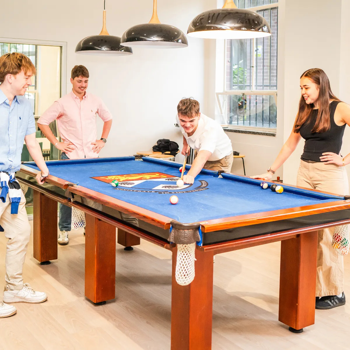 Four young adults playing pool on a blue felt table in a bright room with large windows and overhead lamps.