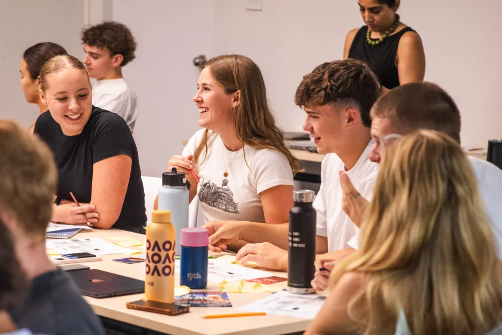 Group of young adults sitting around a table engaged in a lively discussion with notebooks and water bottles.