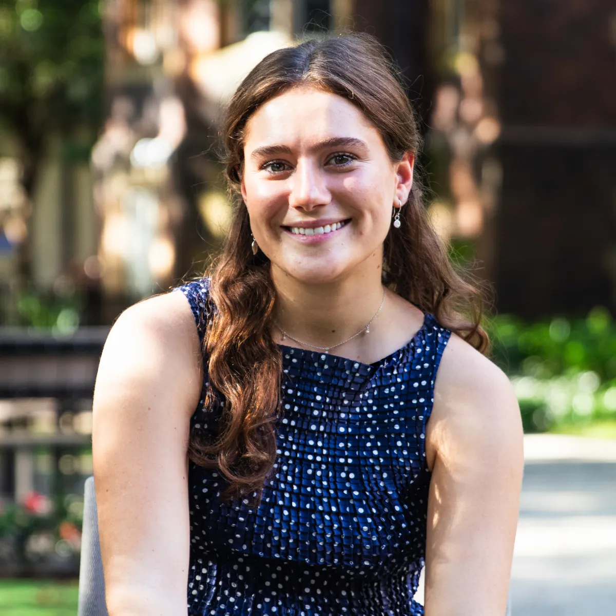 Young woman with long brown hair smiling outdoors, wearing a navy blue patterned sleeveless dress and silver earrings.