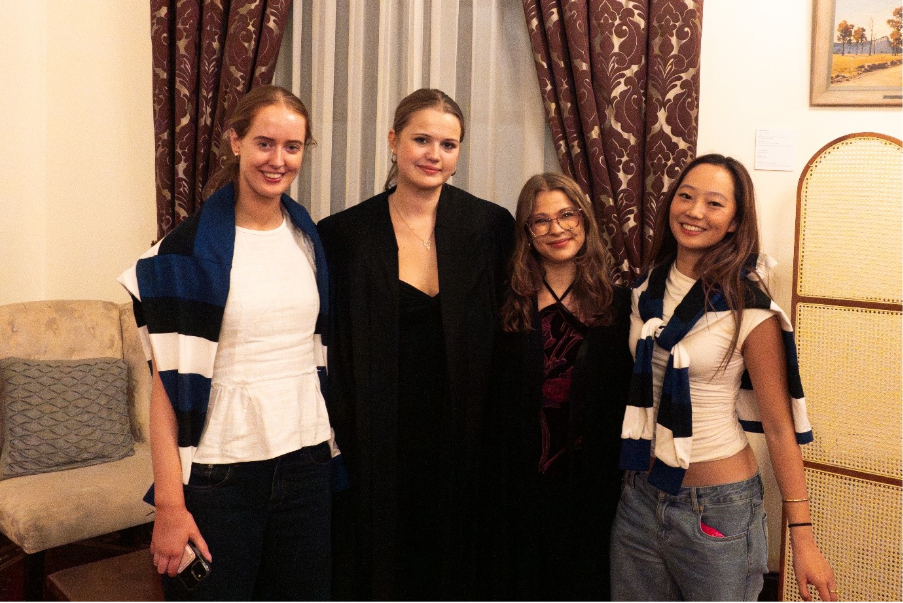 A group of people is standing together, some wearing white shirts, others in black clothing, and a woman in a black gown, near a window with decorative curtains and a piece of art on the wall.AI-generated content may be incorrect.