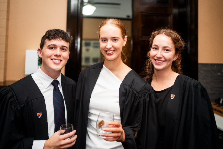 Three individuals dressed in formal attire, with one holding a wine glass, stand in a room with a decorative window and a black desk.AI-generated content may be incorrect.