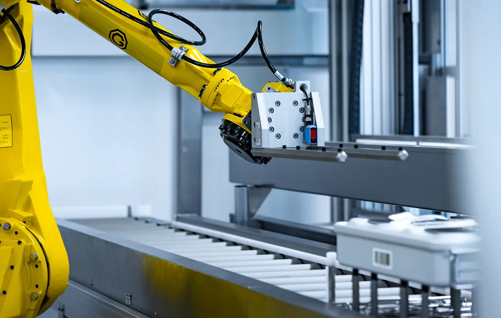 Ein gelber Industrieroboterarm an einem Förderband in einer Fabrik für Metall- und Kunststoffkomponenten.