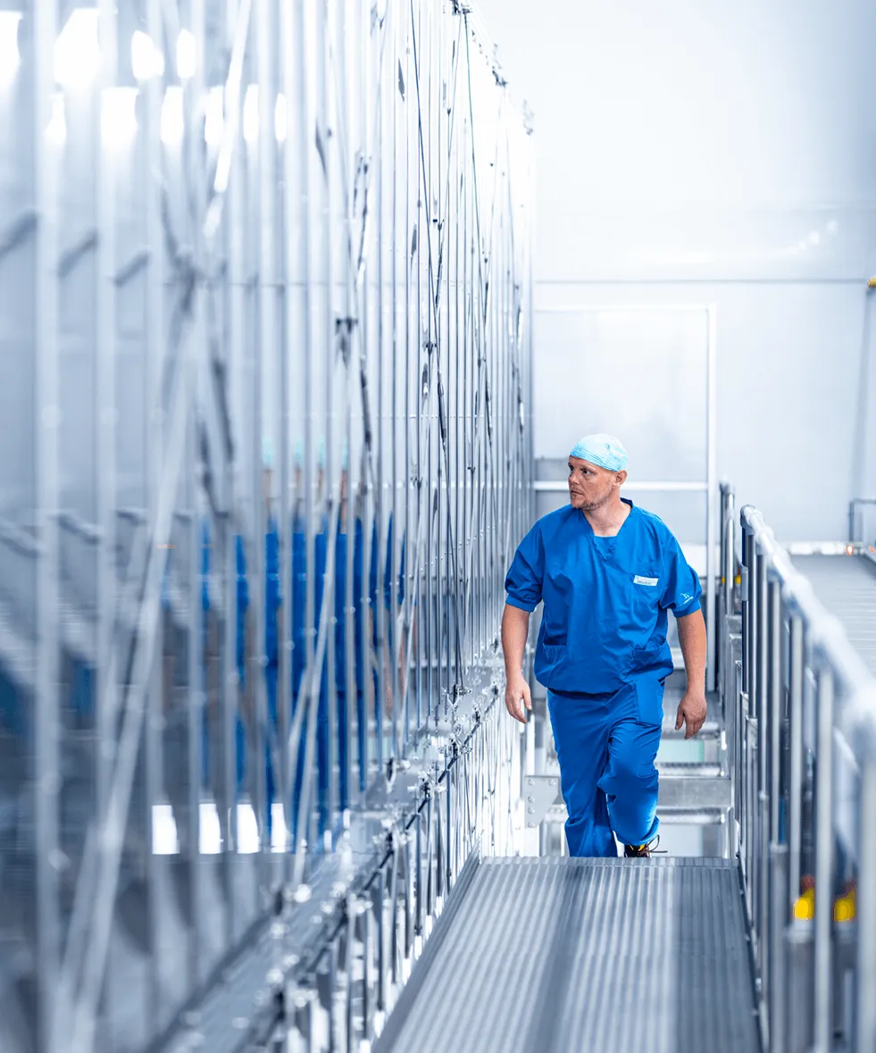 Ein Mann in blauer Arbeitskleidung und einer Mütze geht einen sterilen Gang entlang, an dessen Seite sich eine Metallkonstruktion befindet.