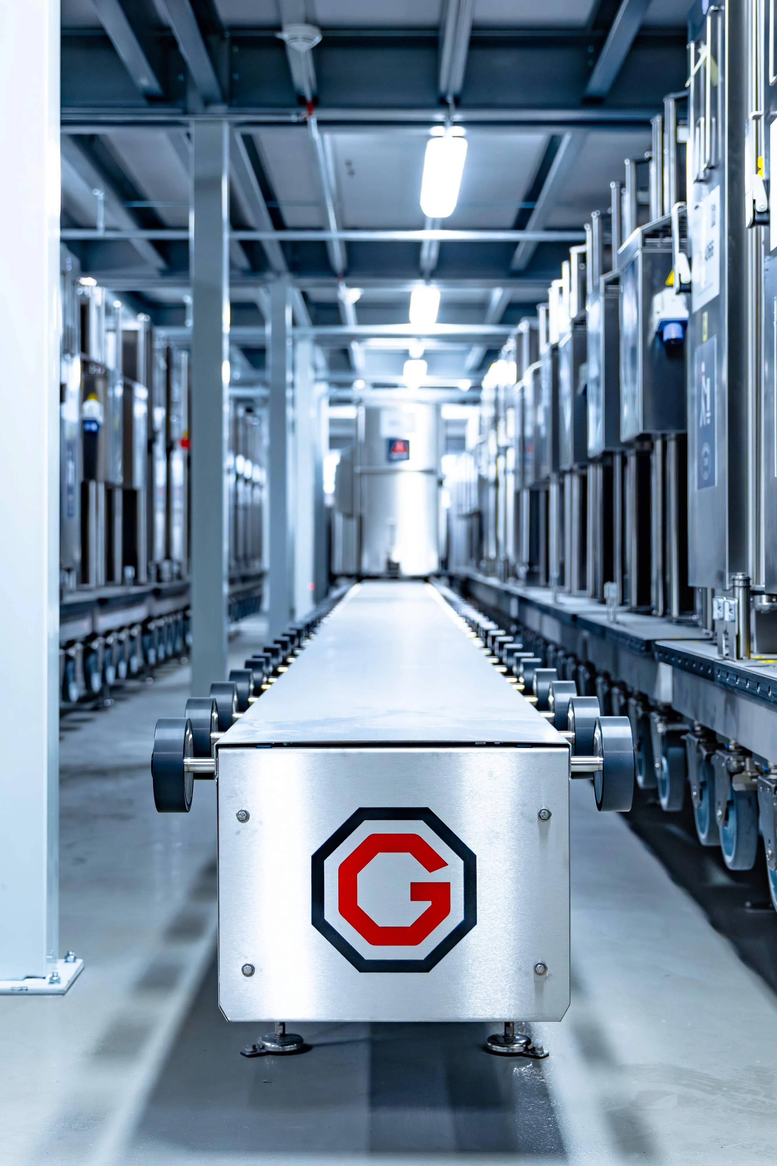 Eine lange Stahlplattform mit schwarz-rotem G-Logo in einer hellen Industrieanlage mit Reihen von Maschinen.