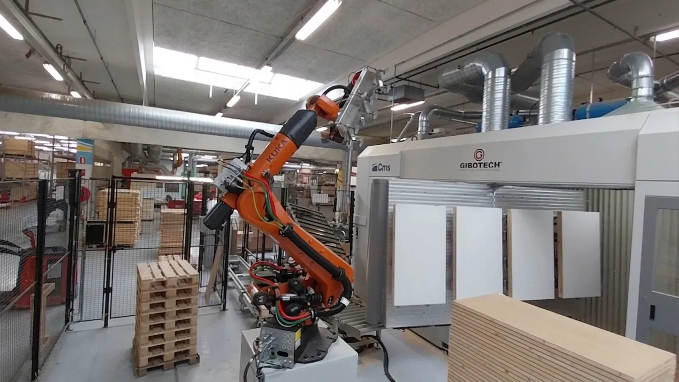 Ein orangefarbener KUKA-Roboterarm transportiert Holzplatten in einer Fabrik mit Industrieanlagen.