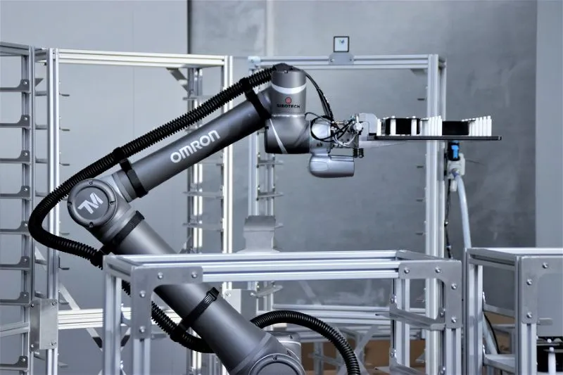 Robothånd med mærket OMRON, der holder en platform med rækker af hvide objekter i et industriel miljø.