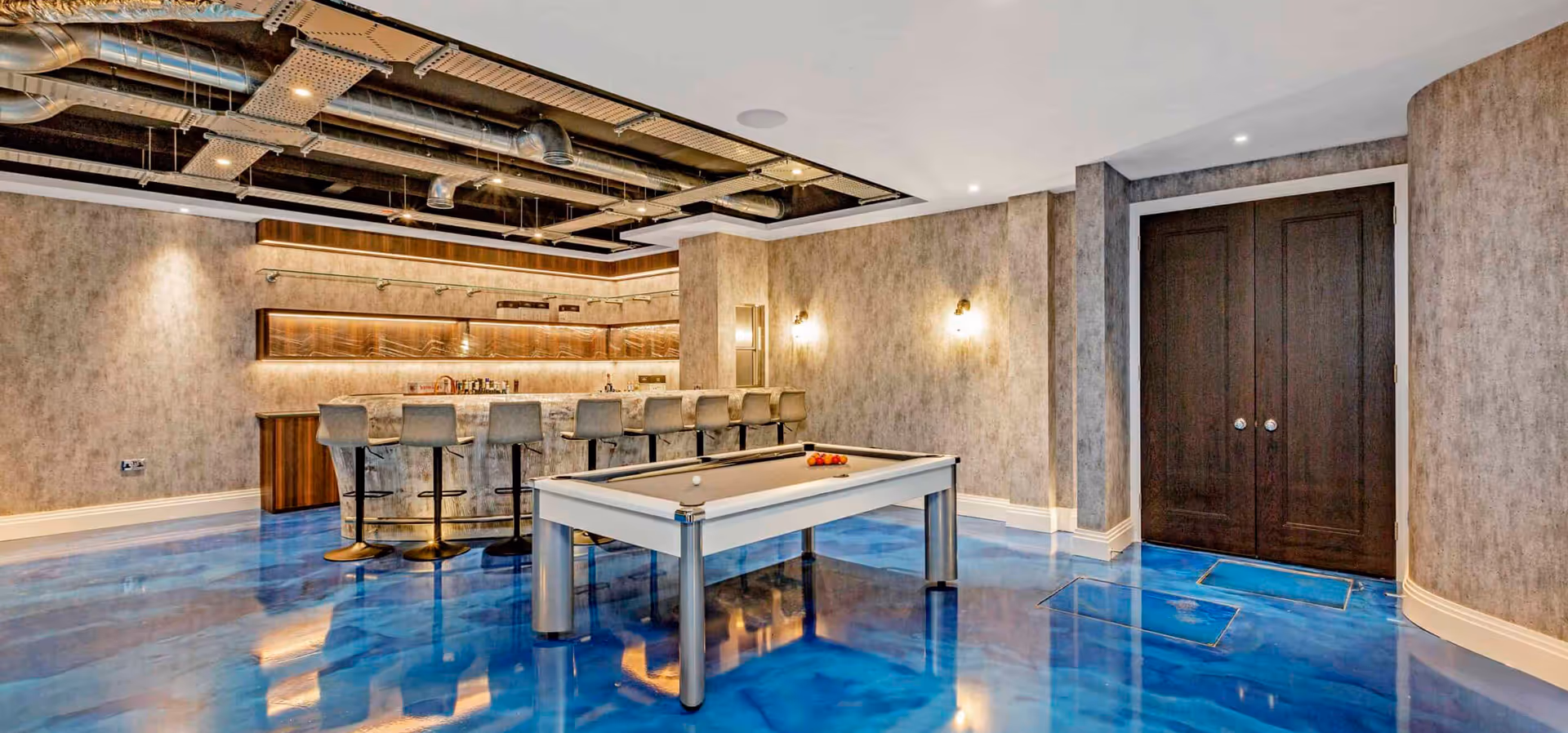 Modern recreational room with a metallic pool table, blue shiny floor, bar with stools, and dark wooden double doors.