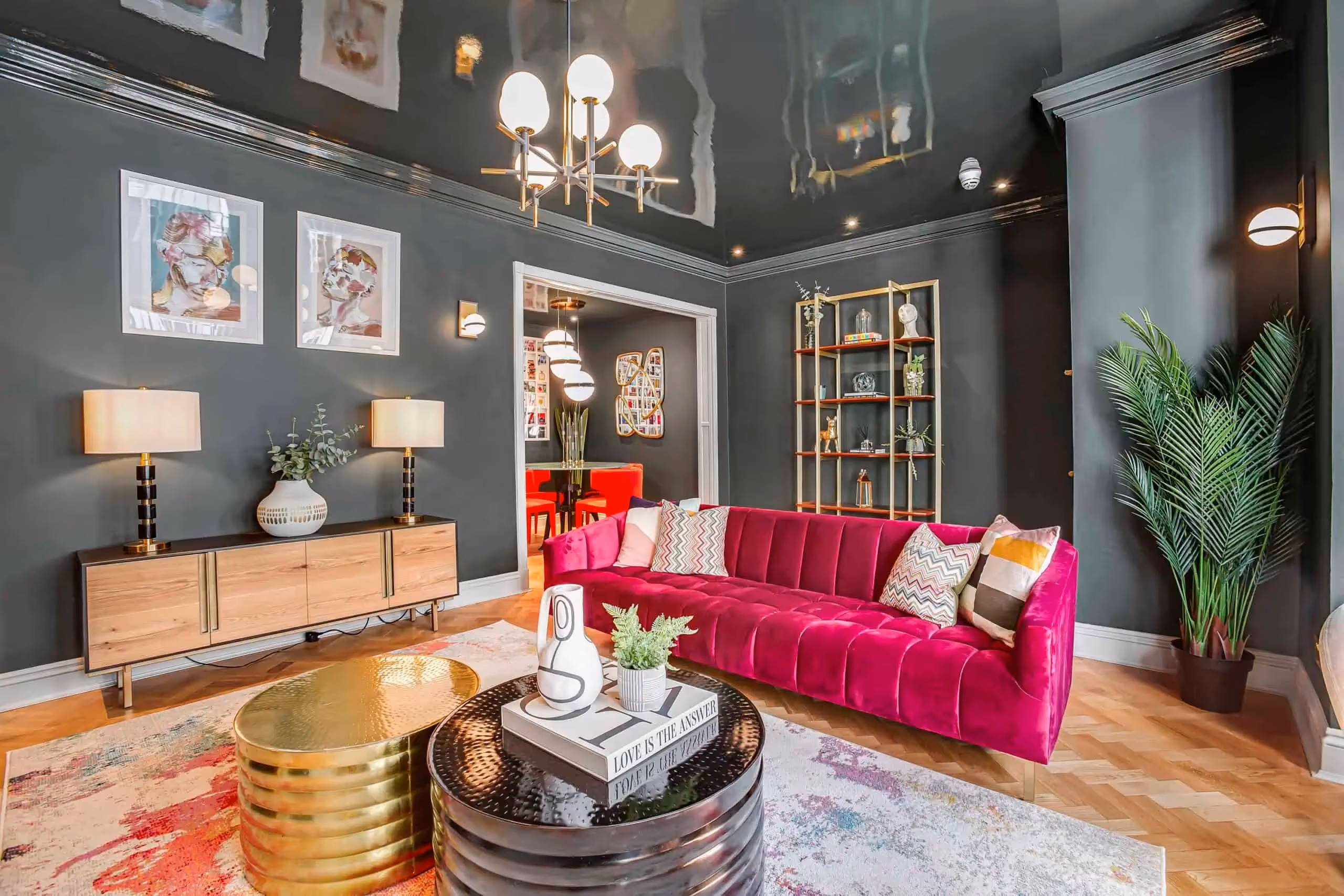 Modern living room with dark grey walls, a bright pink velvet sofa, colourful patterned pillows, gold and black coffee tables, a wooden sideboard with lamps, abstract wall art, and a tall green plant in the corner.