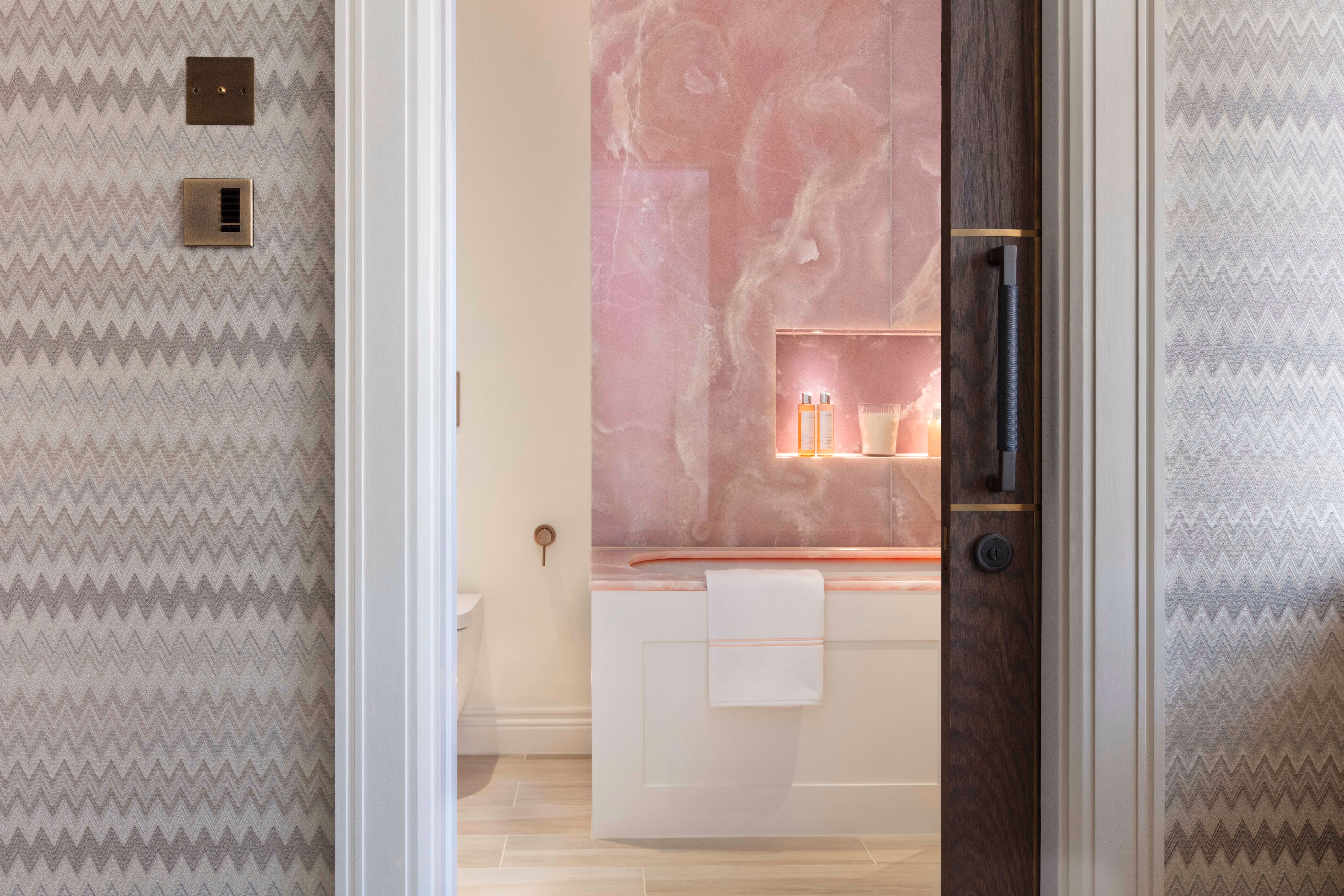 Modern bathroom with pink wall and bathtub, white towel hanging on tub panel, and illuminated shelf with toiletries.