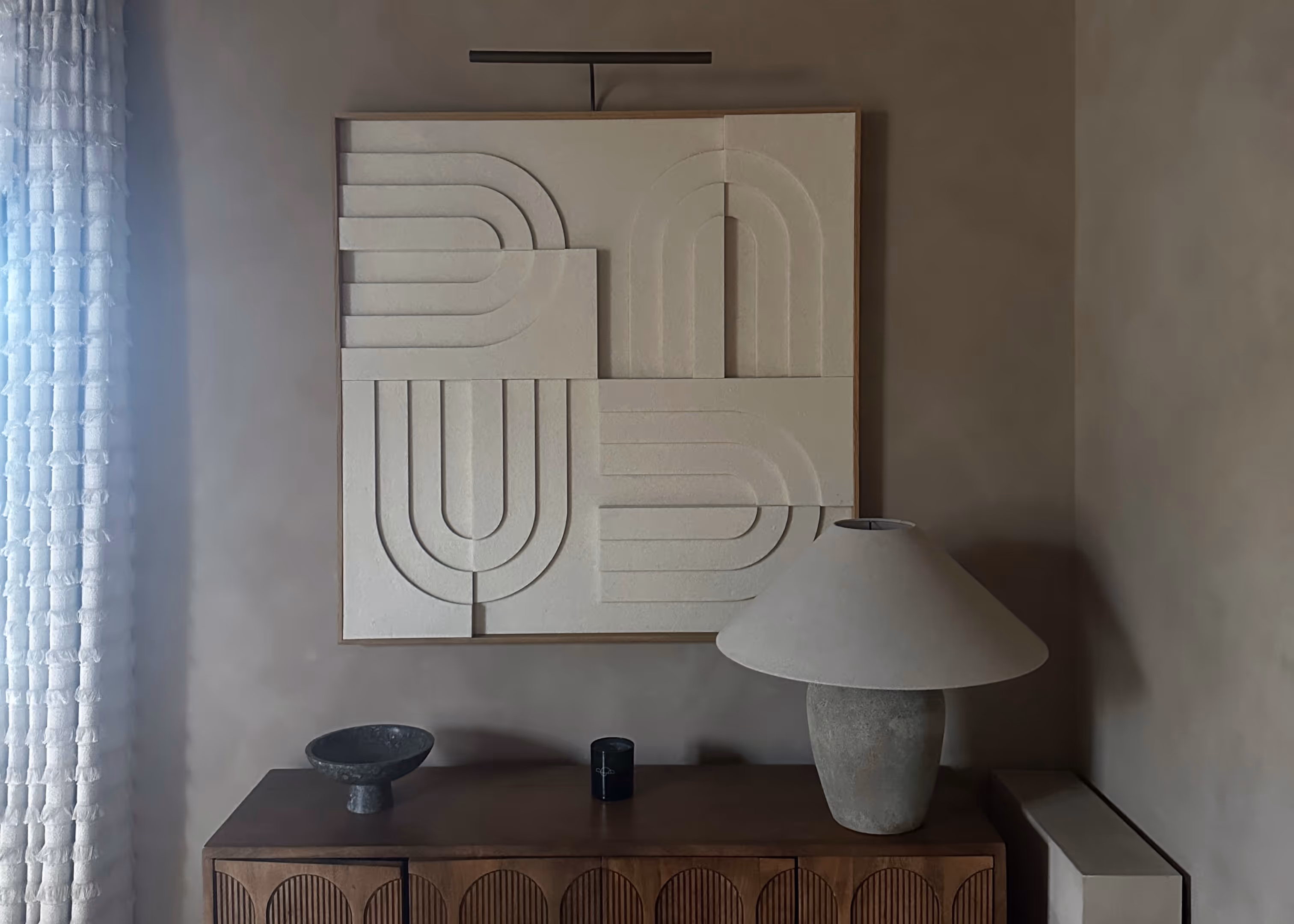 Minimalist interior with geometric wall art, wooden sideboard, ceramic table lamp, and textured white curtain.