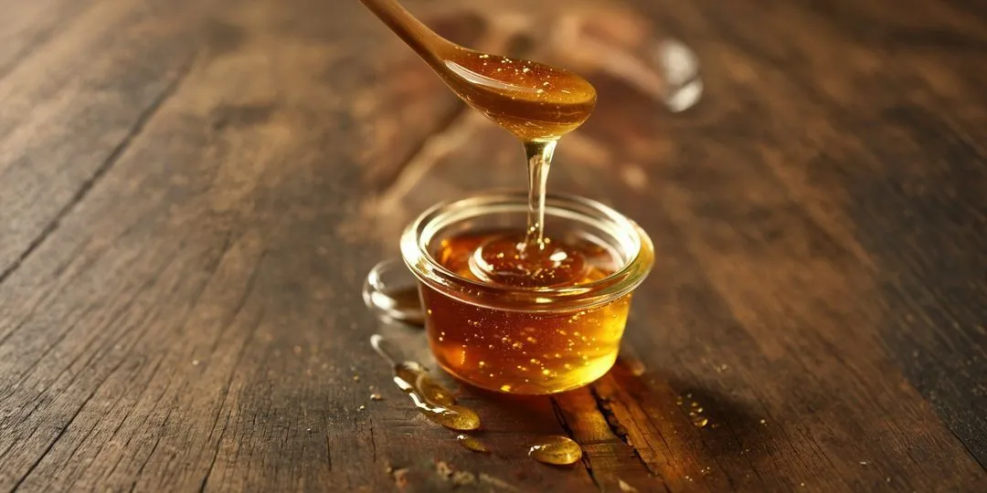 Golden honey dripping from a wooden spoon into a small glass bowl on a rustic wooden surface.