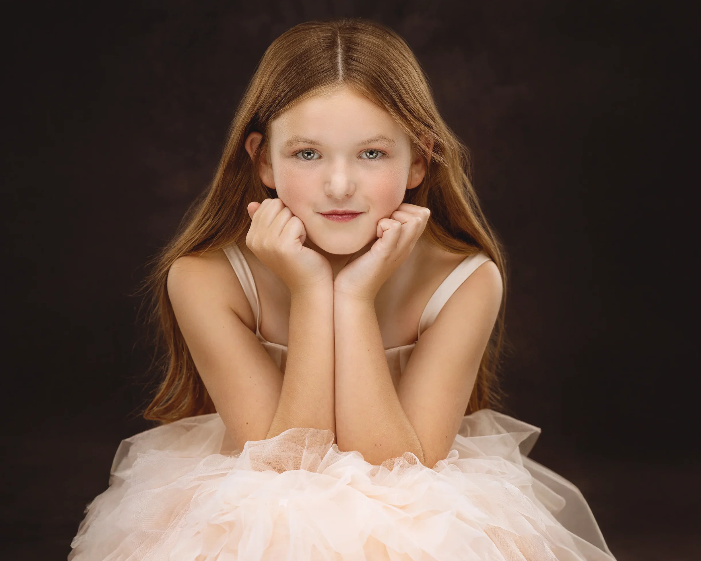Young girl with long brown hair resting her chin on her hands wearing a light pink tulle dress against a dark background.