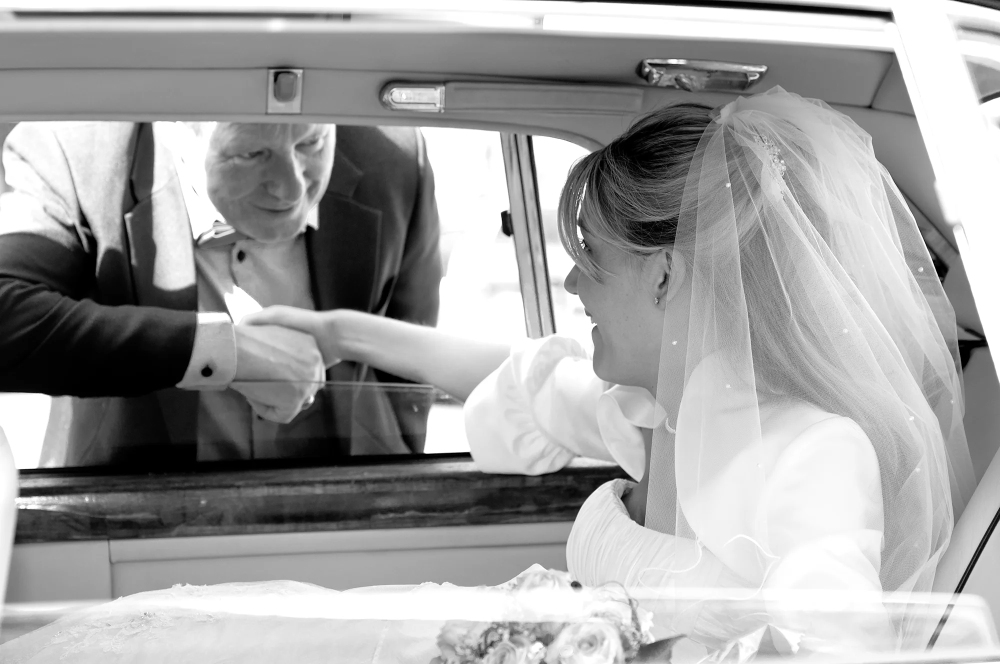 Bride in wedding dress with veil sitting inside a car holding hands with a smiling man outside the car.
