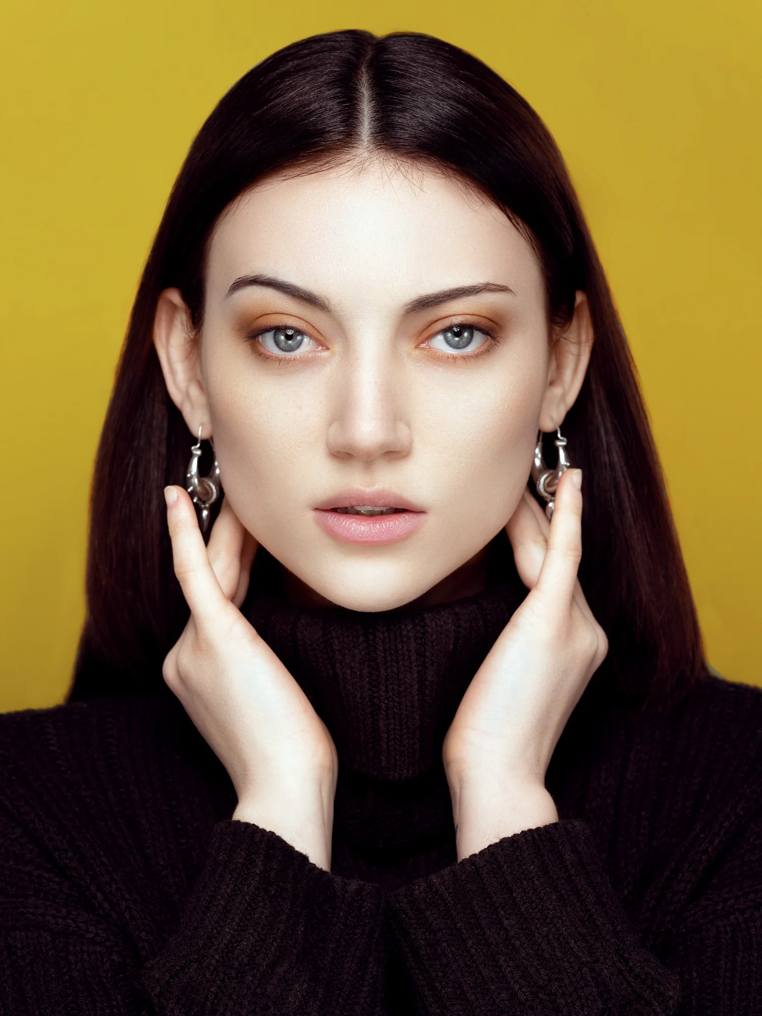 Close-up portrait of a young woman with blue eyes, dark hair, and brown eyeshadow wearing silver earrings and a black turtleneck sweater against a yellow background.
