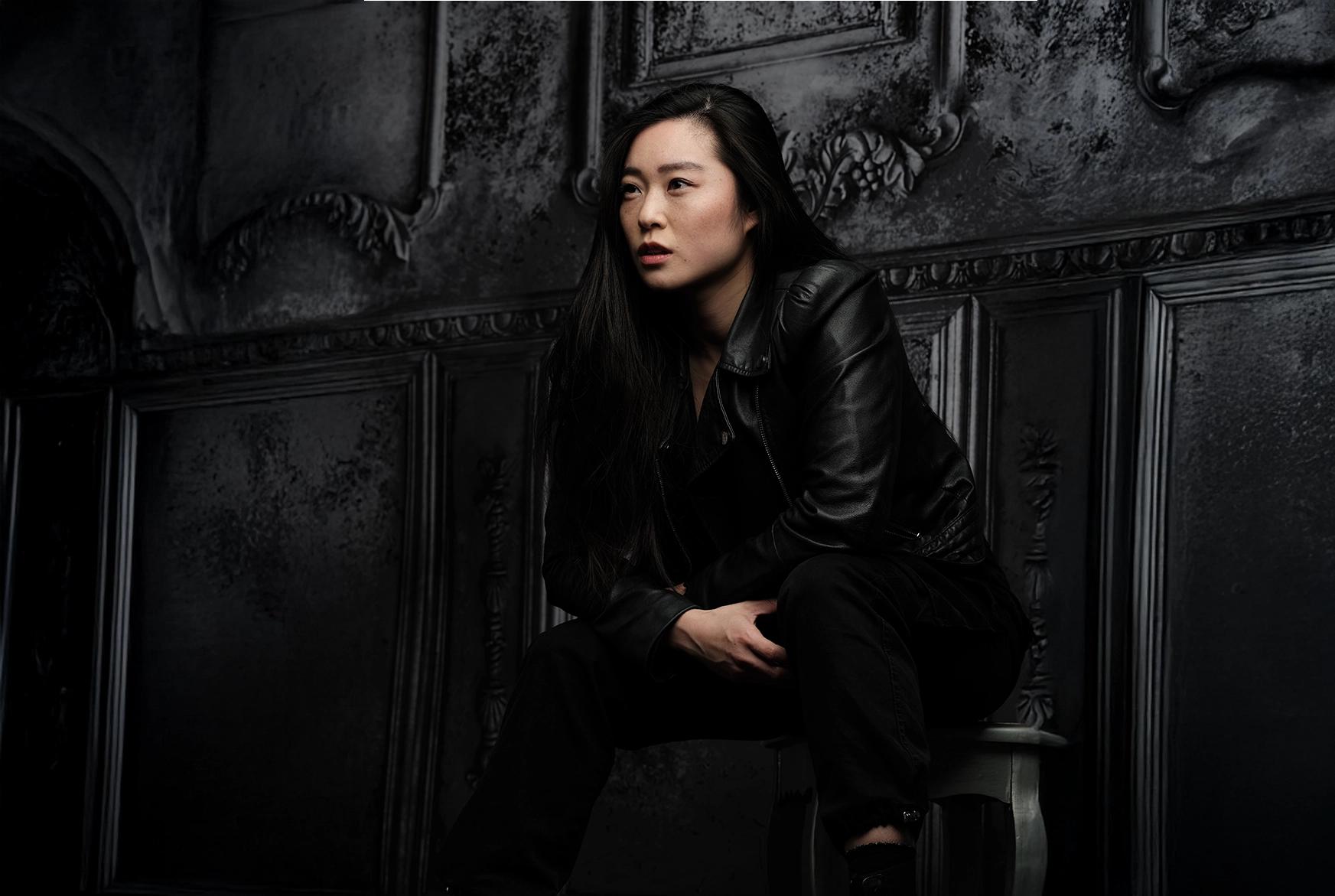 Woman with long dark hair wearing a black leather jacket and black pants sitting on a chair against a dark, ornate wall.