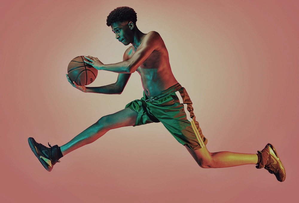 Shirtless young man in green shorts jumping mid-air holding a basketball against a pink background.