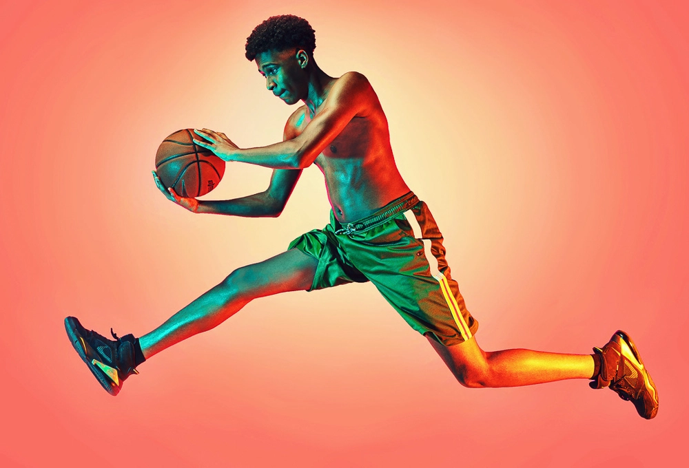 Young shirtless man in green shorts jumping mid-air holding a basketball against an orange background.