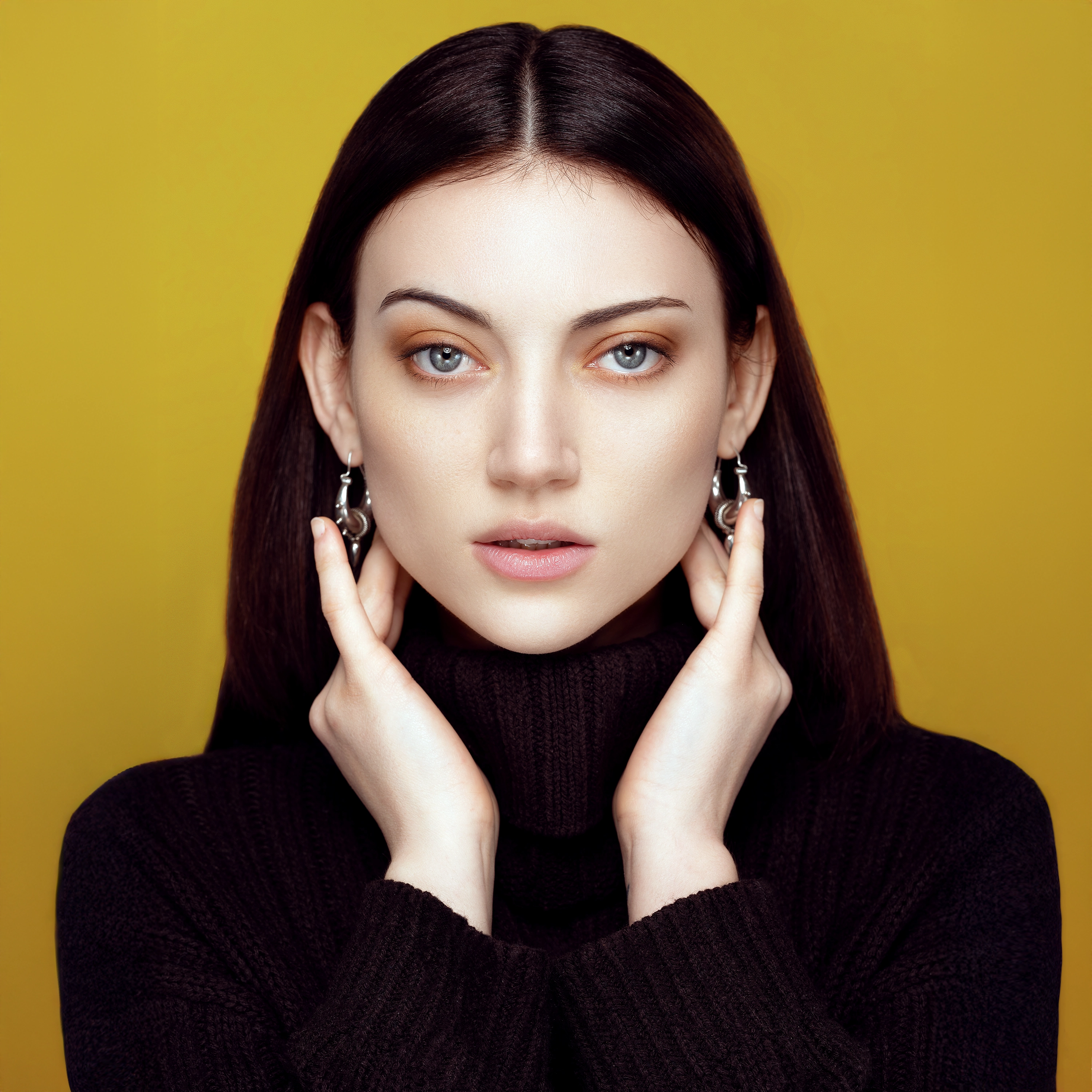 Elyon editing retouching example of a portrait of a woman with straight dark hair wearing silver earrings and a black turtleneck sweater, holding her face with both hands against a yellow background.