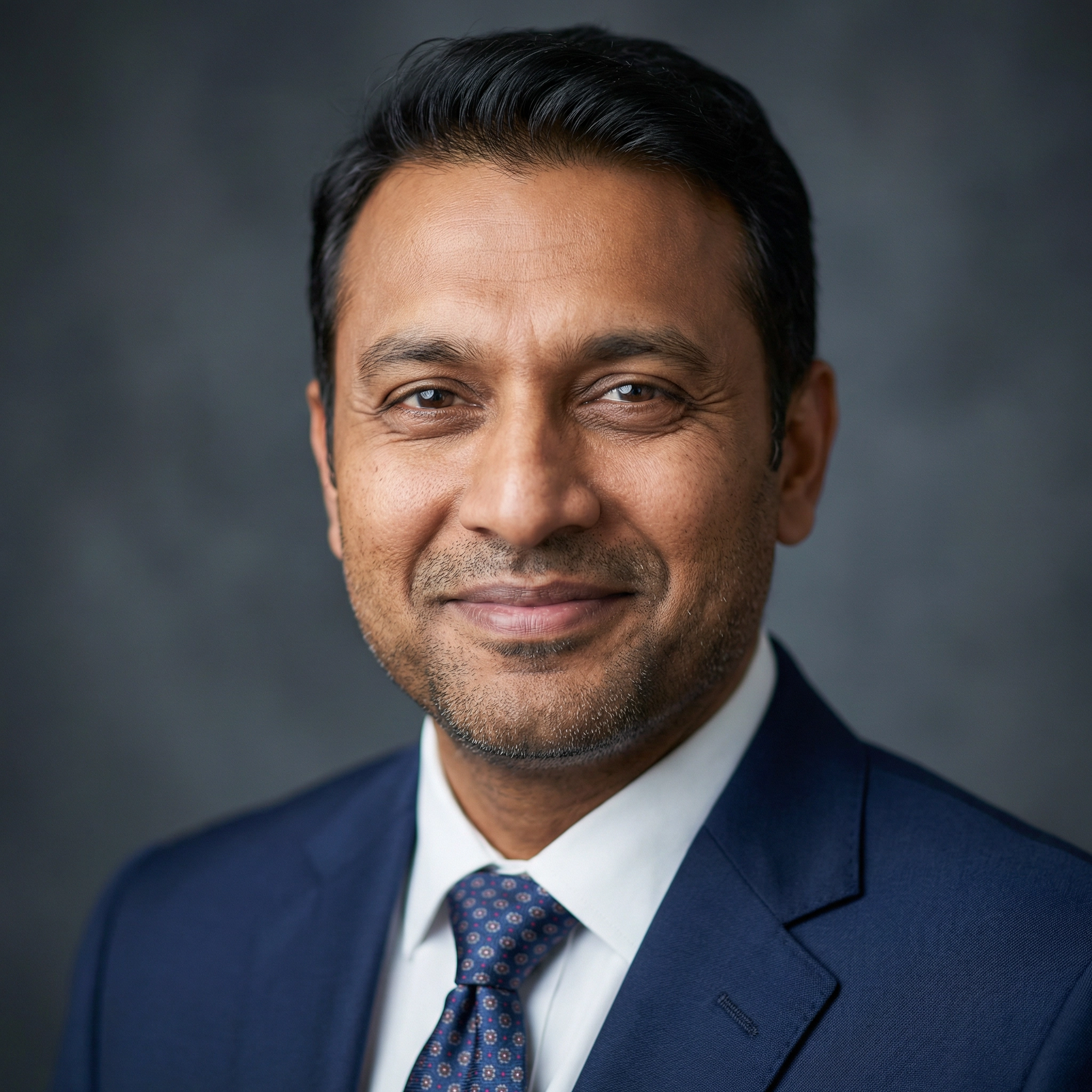 Portrait of a smiling middle-aged man with short black hair, wearing a blue suit jacket, white shirt, and patterned tie against a gray background.