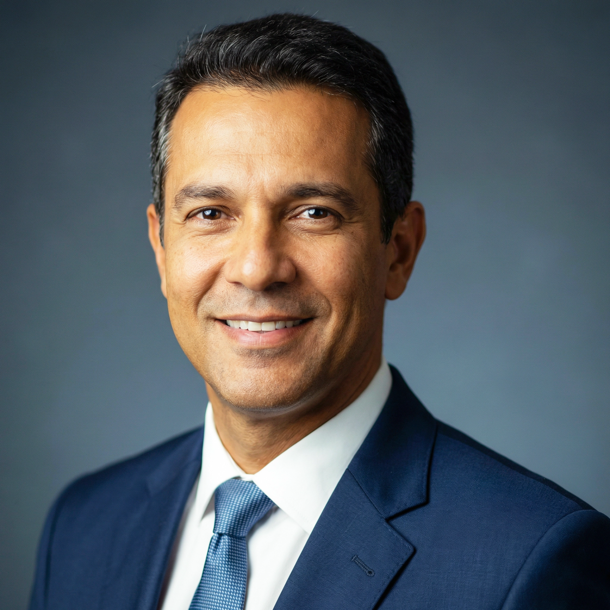 Smiling middle-aged man with short dark hair wearing a blue suit, white shirt, and blue tie against a plain gray background.