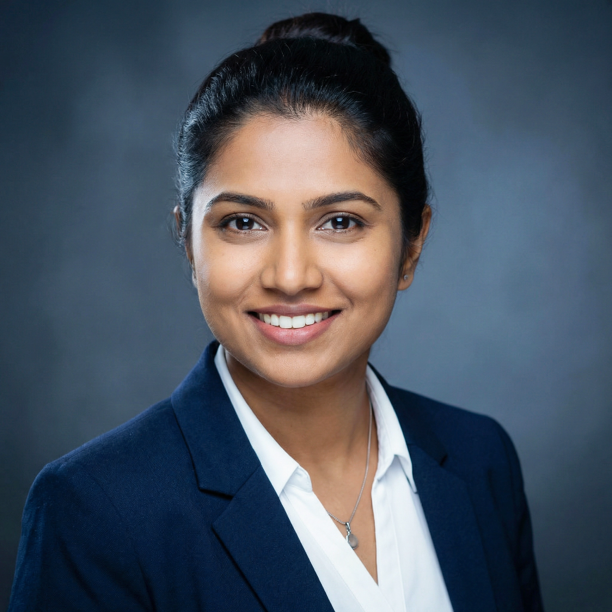 Smiling woman with dark hair in a bun, wearing a navy blazer and white shirt, against a gray background.