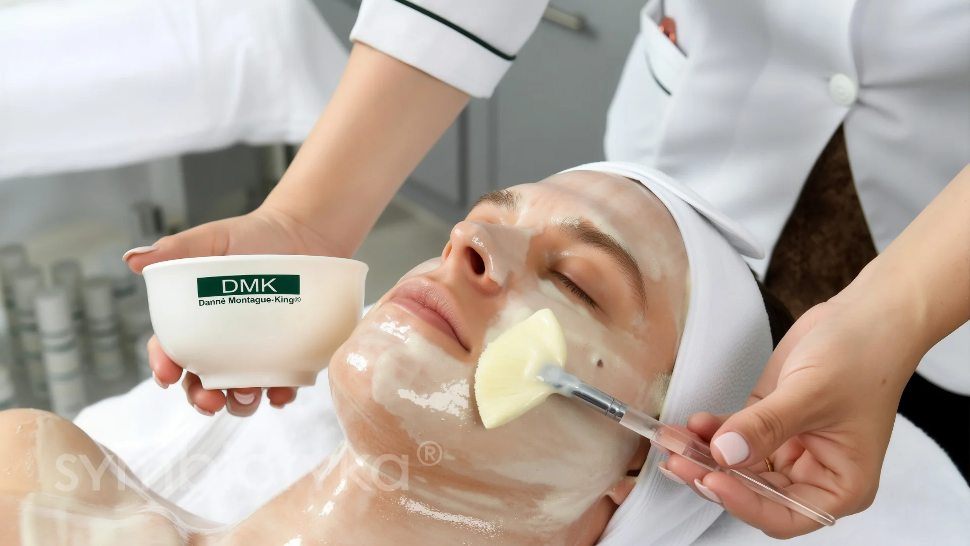 Woman receiving a facial treatment with a white cream mask applied with a brush by a beautician.