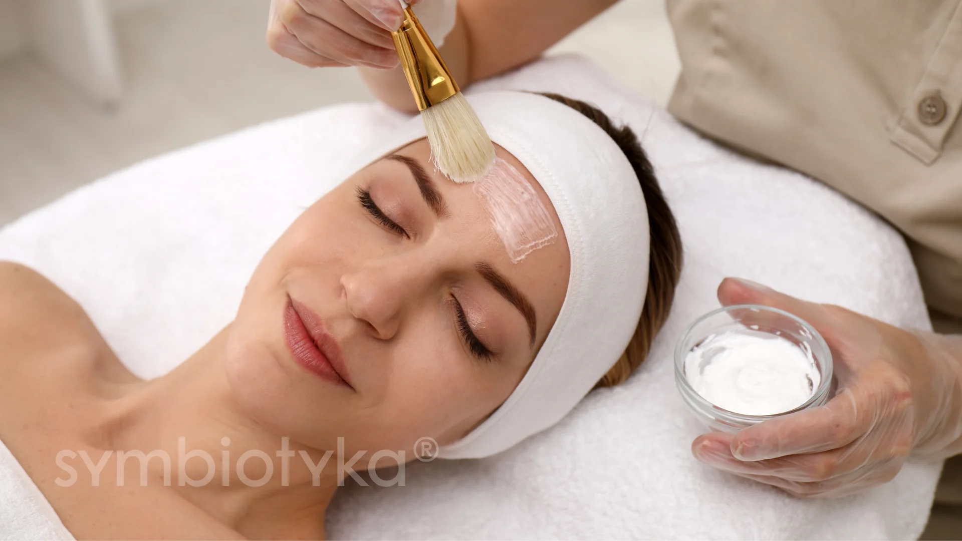 Woman with eyes closed wearing a white headband receiving a facial cream application on her forehead with a brush at a spa.