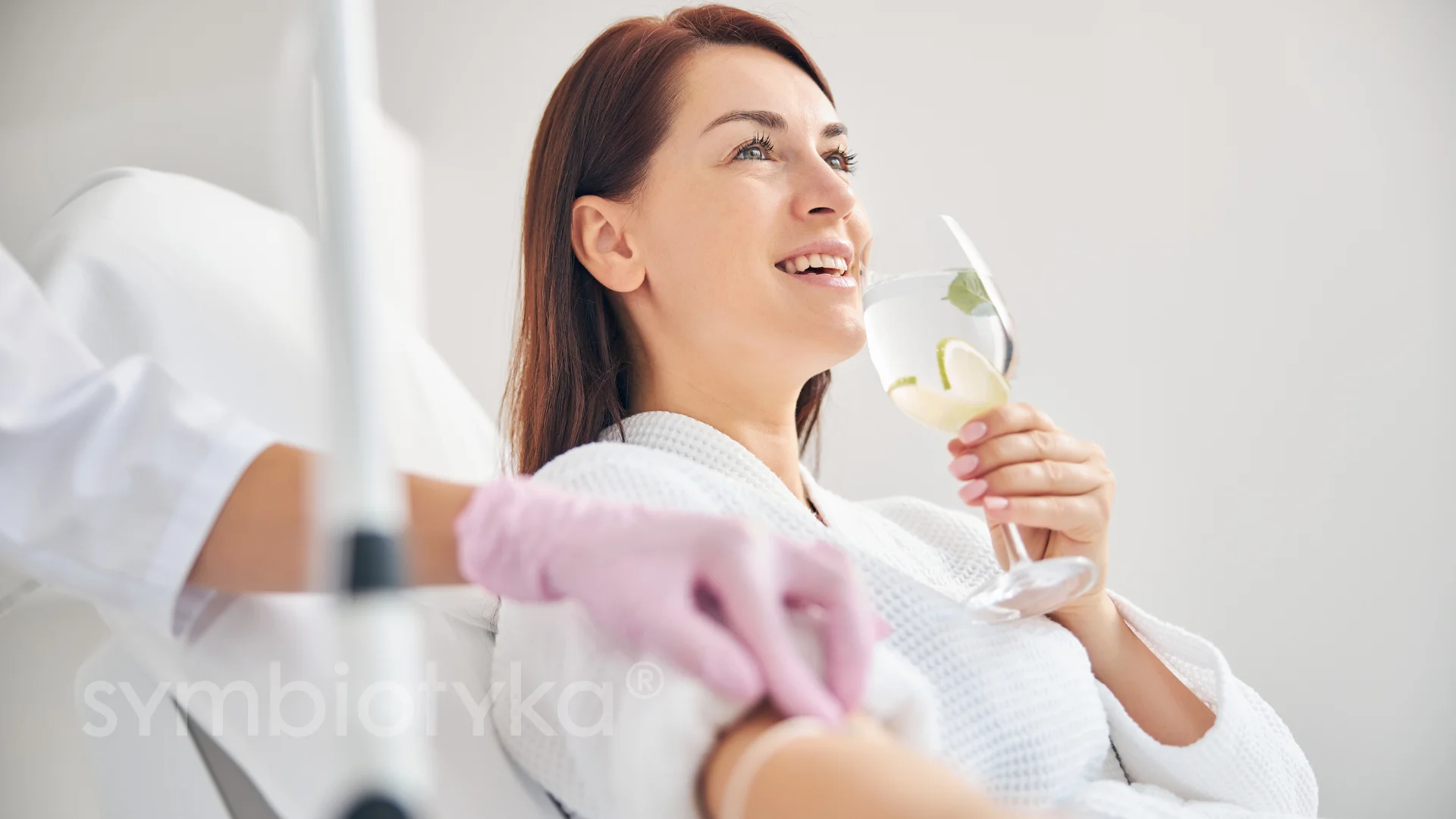 Smiling woman in a white robe holding a glass with lemon water while receiving an IV drip.