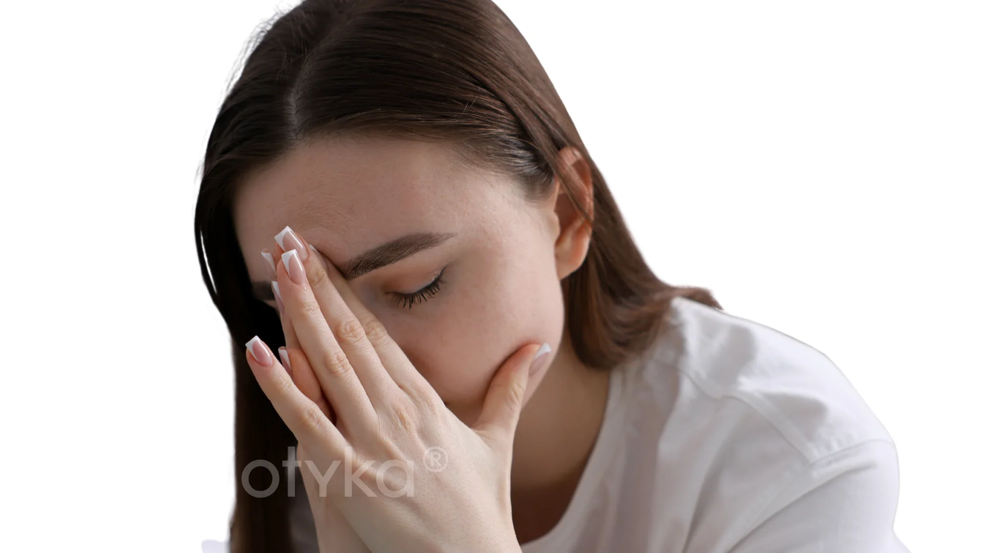 Young woman in white shirt with long brown hair covering her face with her hands, eyes closed, appearing distressed.