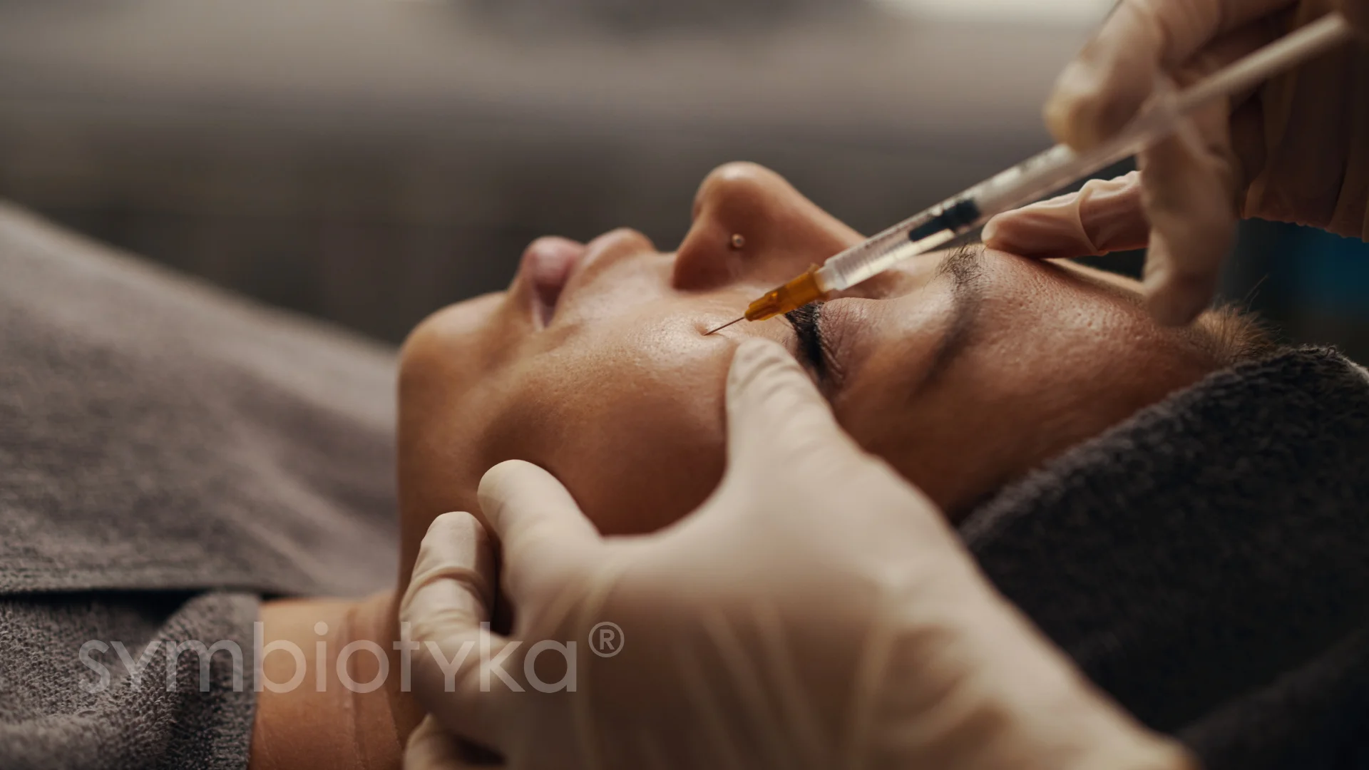 Close-up of a gloved hand injecting a substance with a syringe into the skin near a person's closed eye.