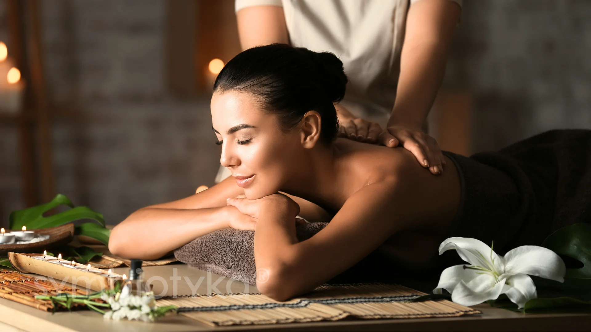 A woman with eyes closed receiving a relaxing back massage in a spa setting with lit candles and white flowers nearby.